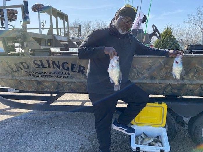 a man standing beside a boat and holding 2 white crappie 