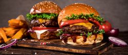 Two gourmet burgers with beef patties, cheese, lettuce, and tomato on sesame seed buns served on wooden cutting board with sweet potato fries