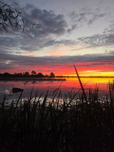 Sunset over lake with vibrant orange and pink sky reflected in calm water, silhouetted trees in foreground, Elma New York