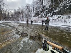 Anglers explore the icy river, surrounded by a serene winter landscape of snow-covered trees and frozen waterfalls.