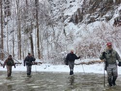 A group of intrepid anglers navigates the snow-covered, partially frozen stream, undeterred by the wintry conditions as they pursue their next catch in this serene, forested landscape.