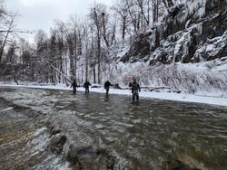 Hikers trek through the snow-covered forest, navigating the icy stream with caution as towering cliffs loom in the distance, creating a breathtaking winter landscape.