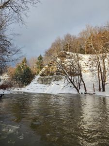 Tranquil winter wonderland: A serene snow-covered scene with a still body of water reflecting the surrounding bare trees and rocky outcrop.