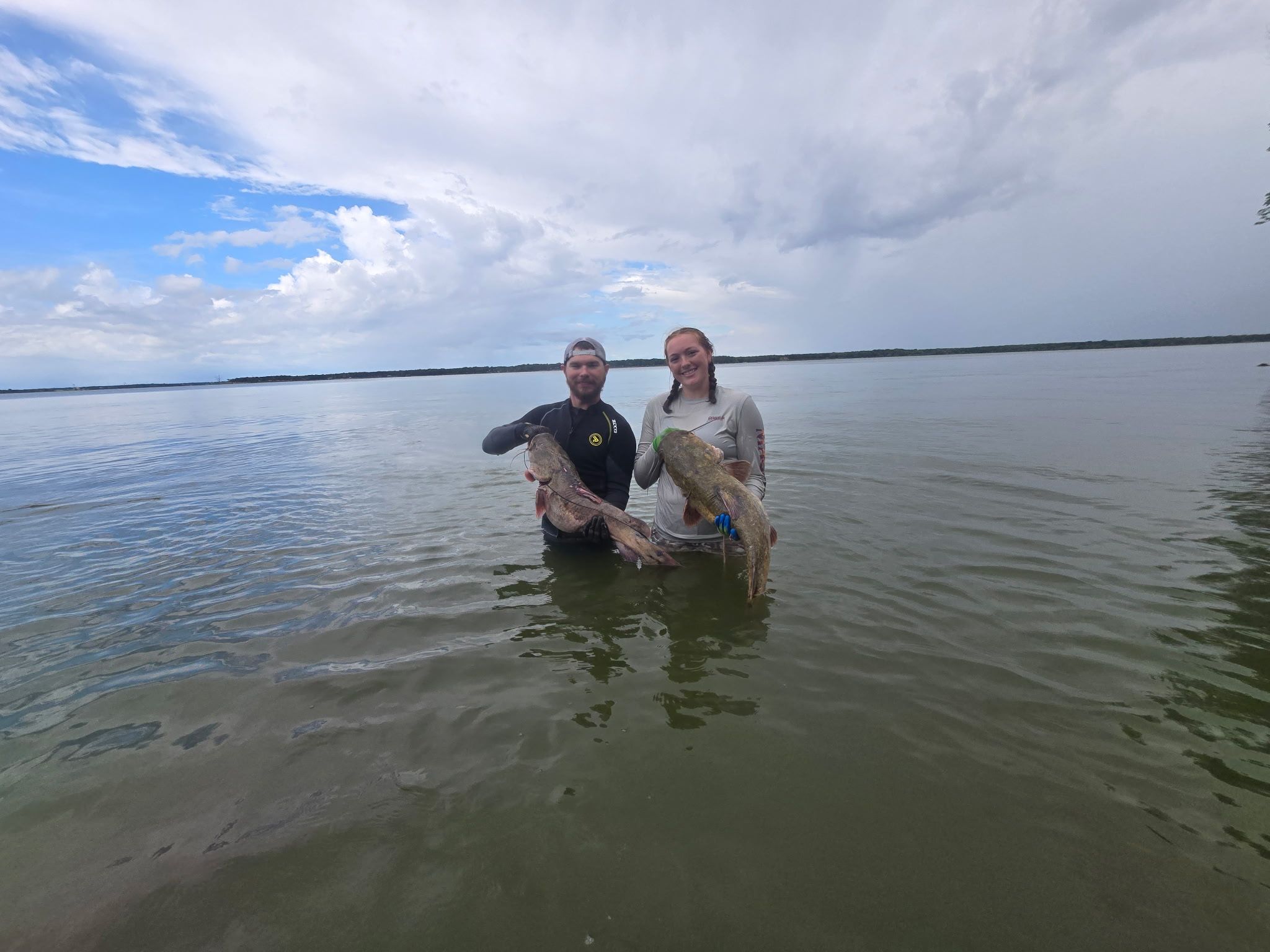 Two anglers standing in shallow water each holding a large flathead catfish
