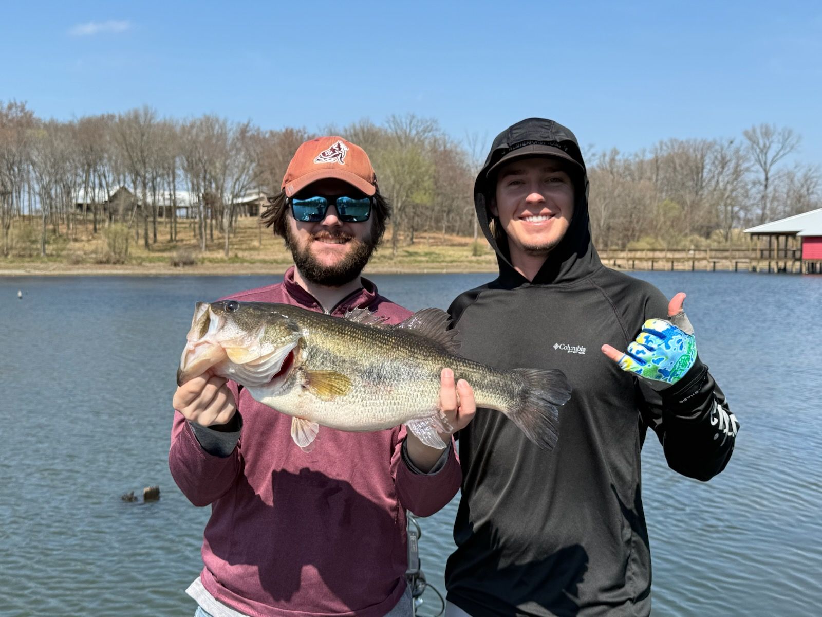 Two anglers holding a freshly caught bass by a lake on a clear day