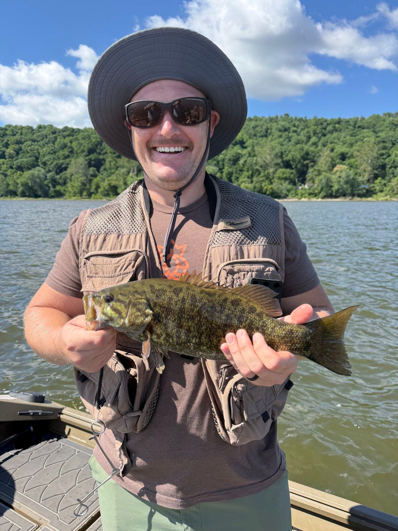Angler holding freshly caught smallmouth bass on boat with forested shoreline in background