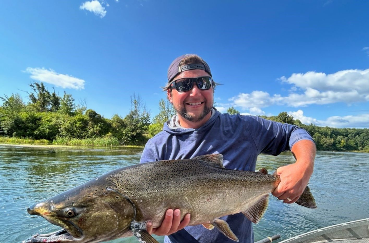 Angler holding a large Chinook salmon on a boat in a river surrounded by green trees