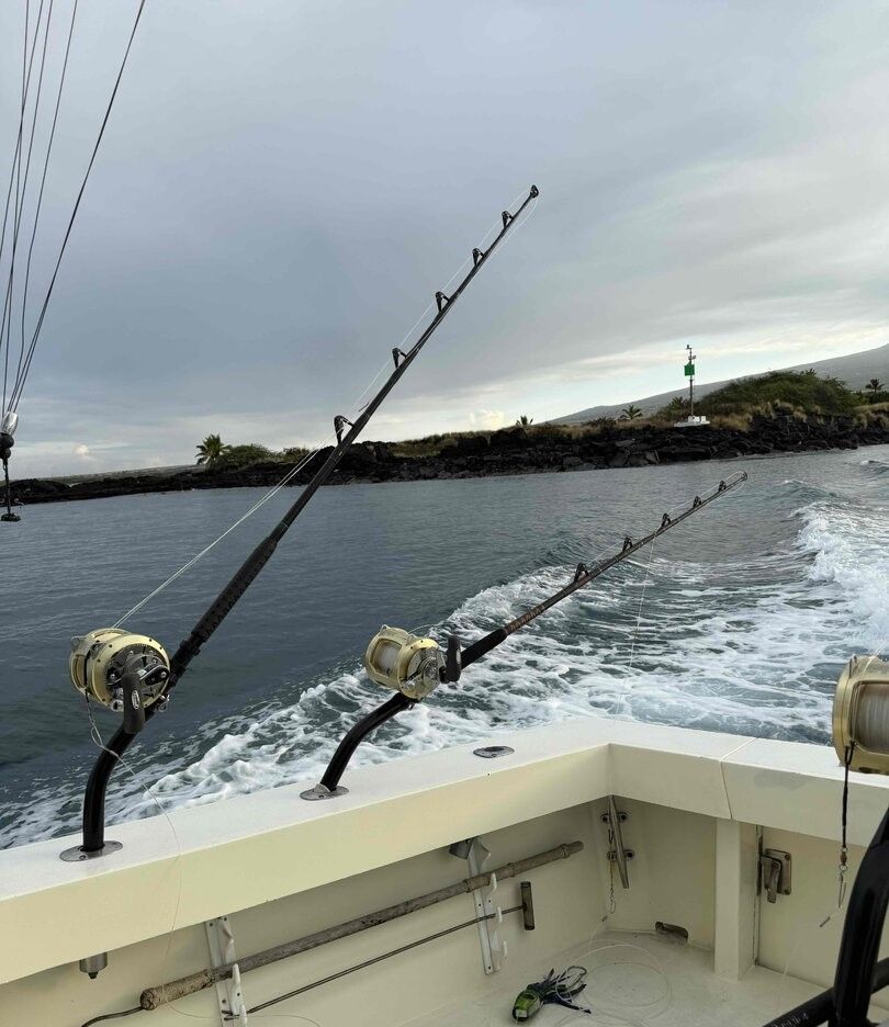 Fishing boat with multiple rods set up for deep sea fishing, creating wake in ocean water near rocky coastline with lighthouse