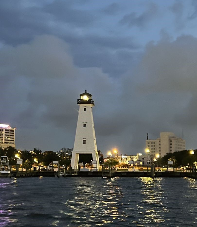 White lighthouse illuminated at dusk with light beam shining, waterfront buildings and boats visible in background