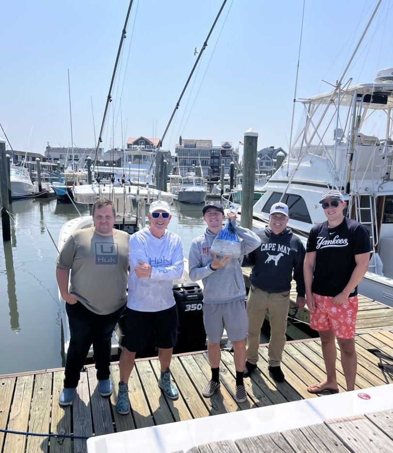 Group of five anglers standing on marina dock with fishing boats and harbor in background