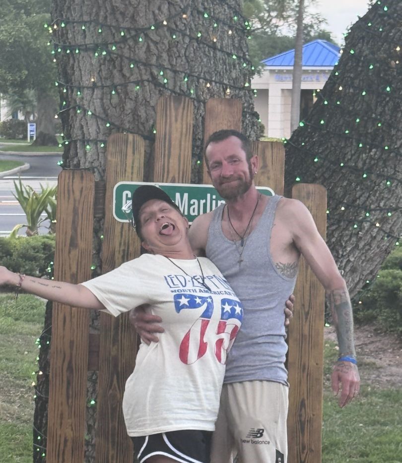 Two people posing together outdoors near wooden fence with string lights in trees