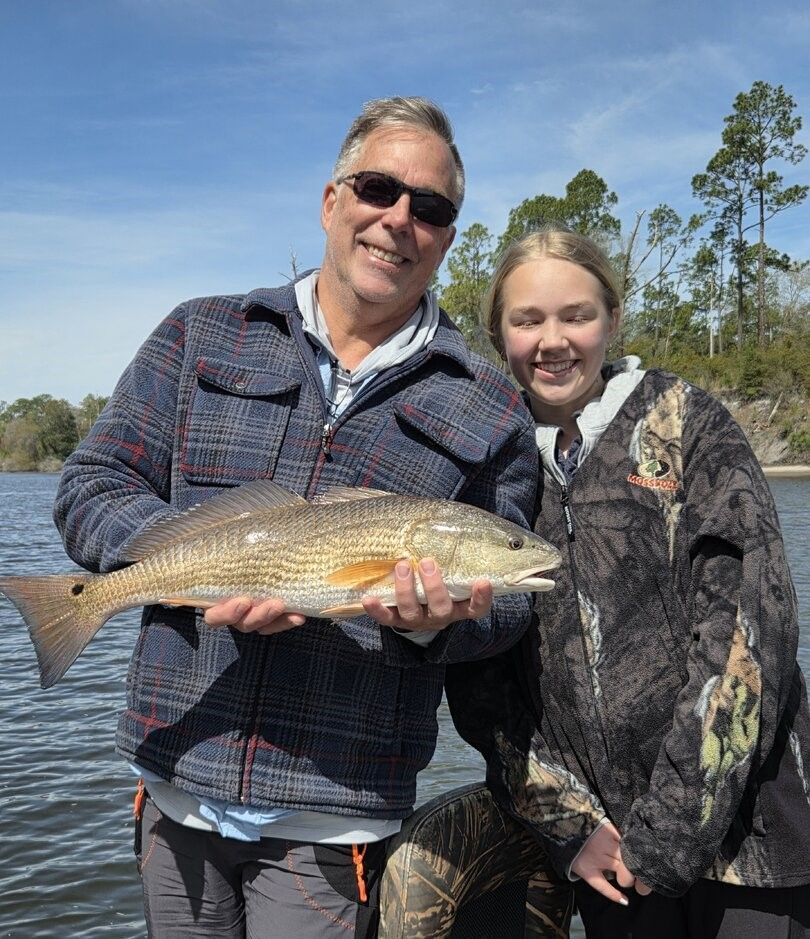 Two people holding a freshly caught redfish on a boat with trees and water in the background