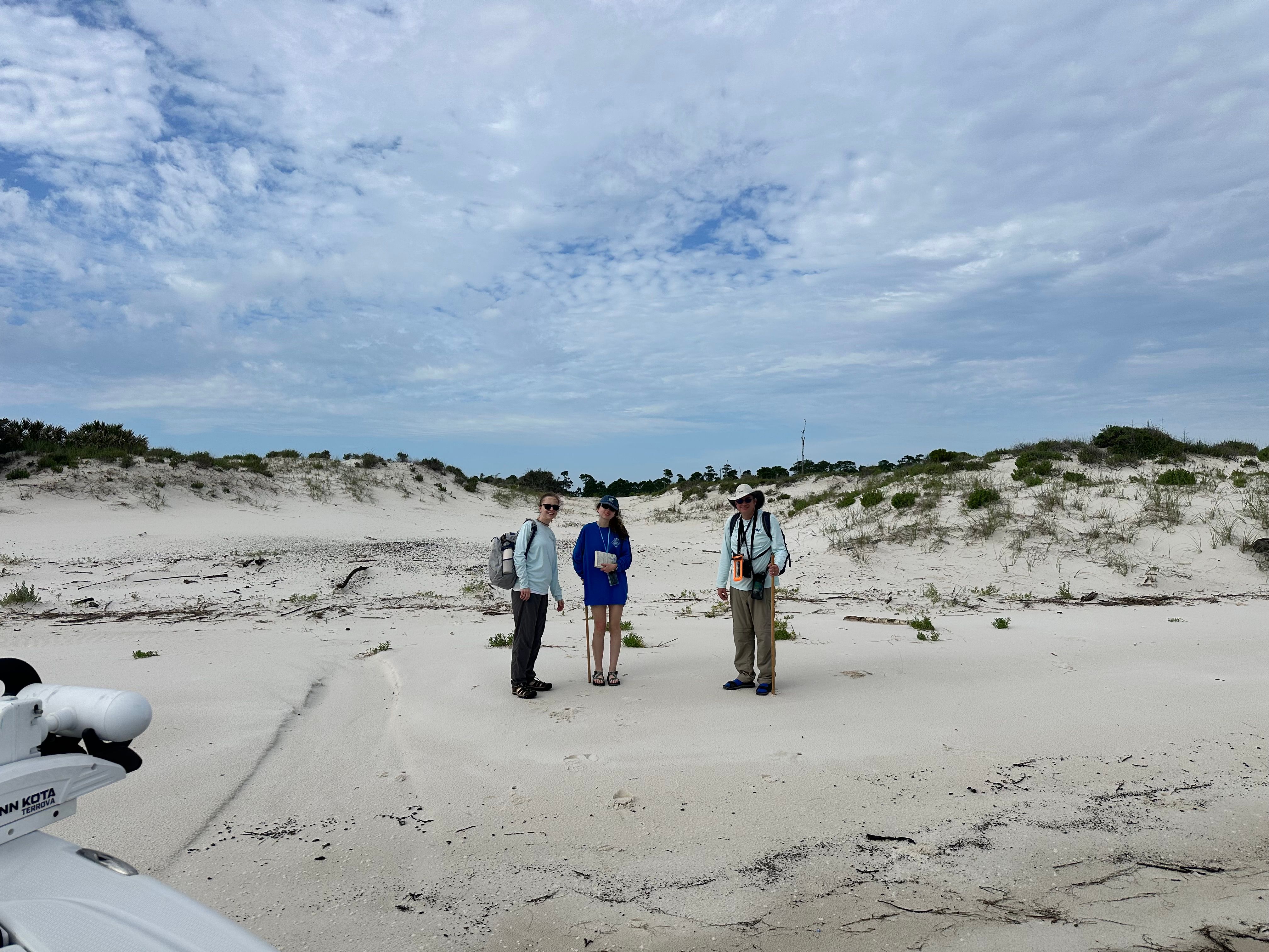 Three people standing on sandy beach with dunes and sparse vegetation under cloudy sky