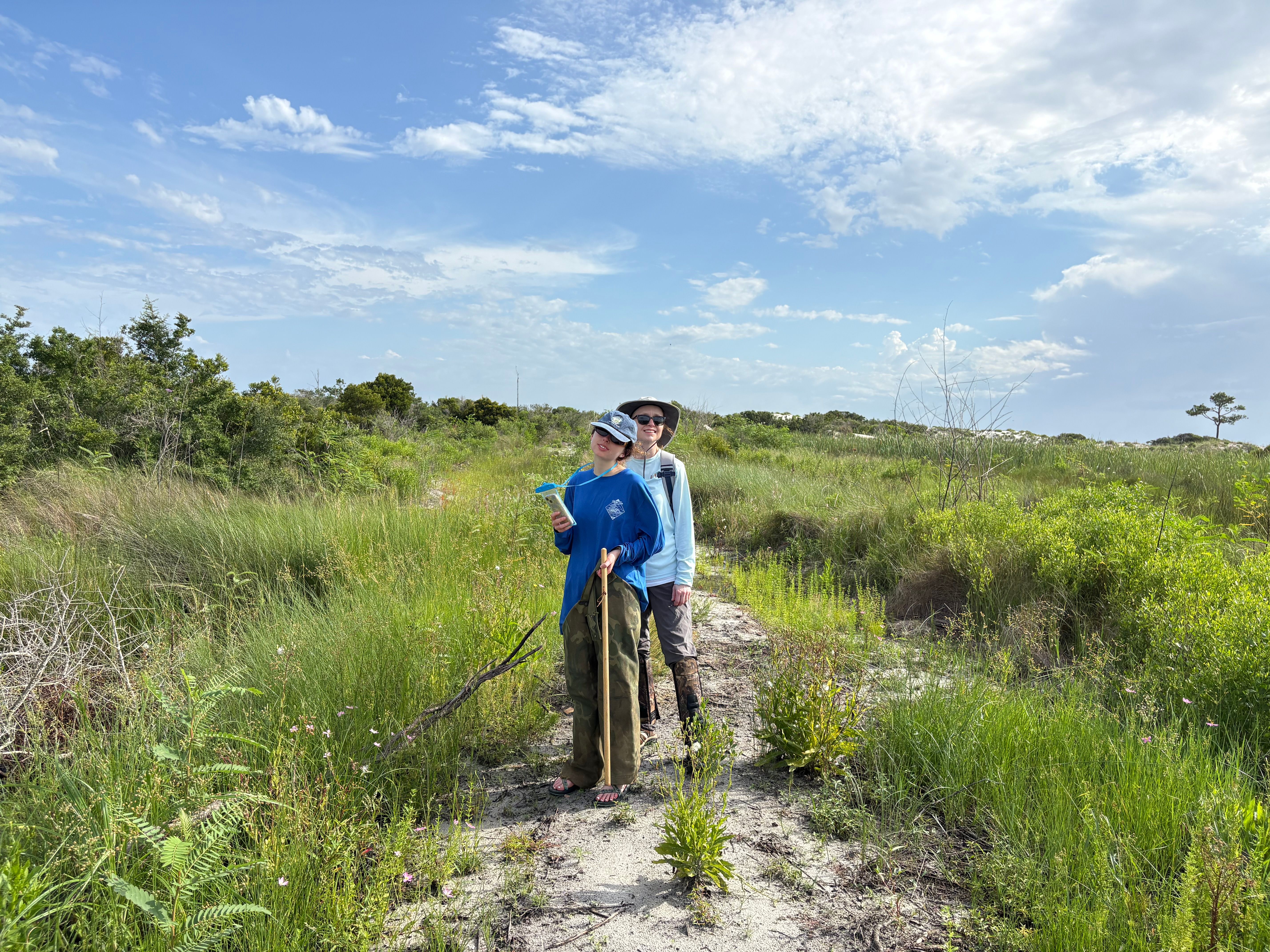 Two hikers standing on a dirt trail surrounded by lush green vegetation and bushes