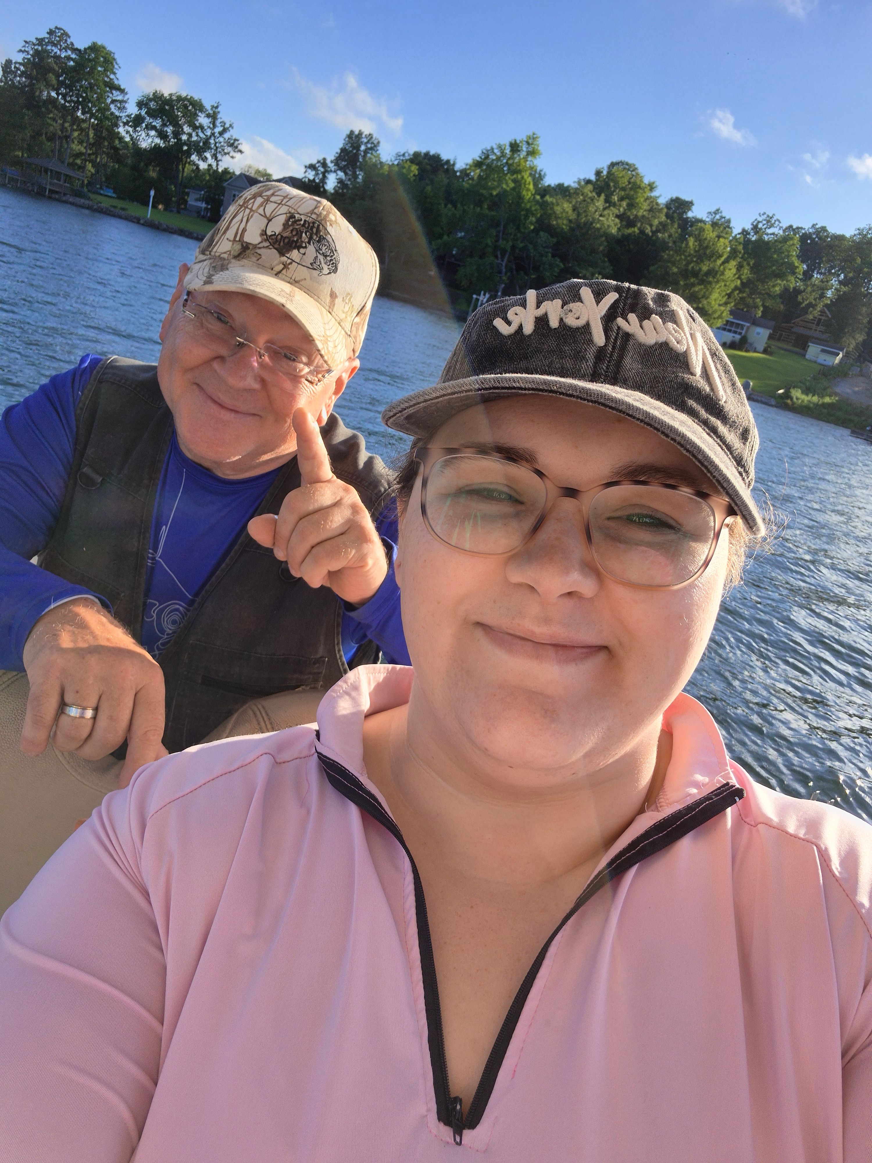Two people standing together outdoors near a lake with trees and blue sky in background