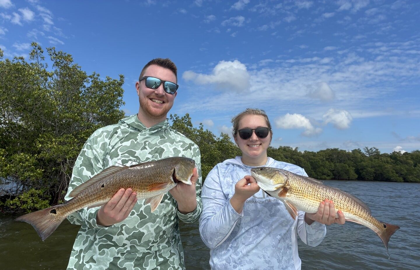 Two anglers displaying freshly caught redfish by waterside with trees and blue sky