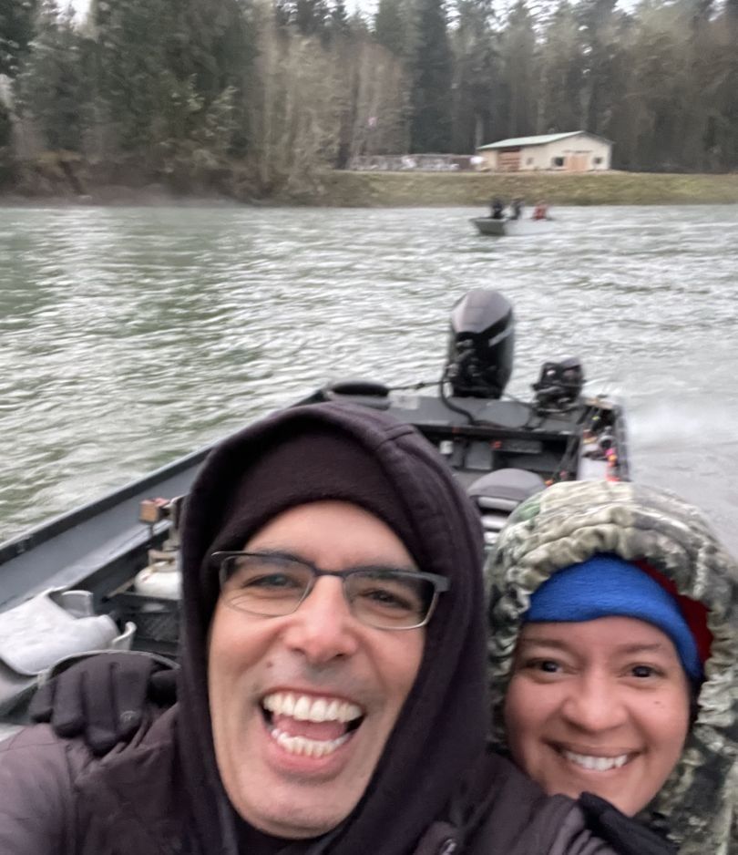 Two anglers smiling on fishing boat with motor and equipment on river with trees in background
