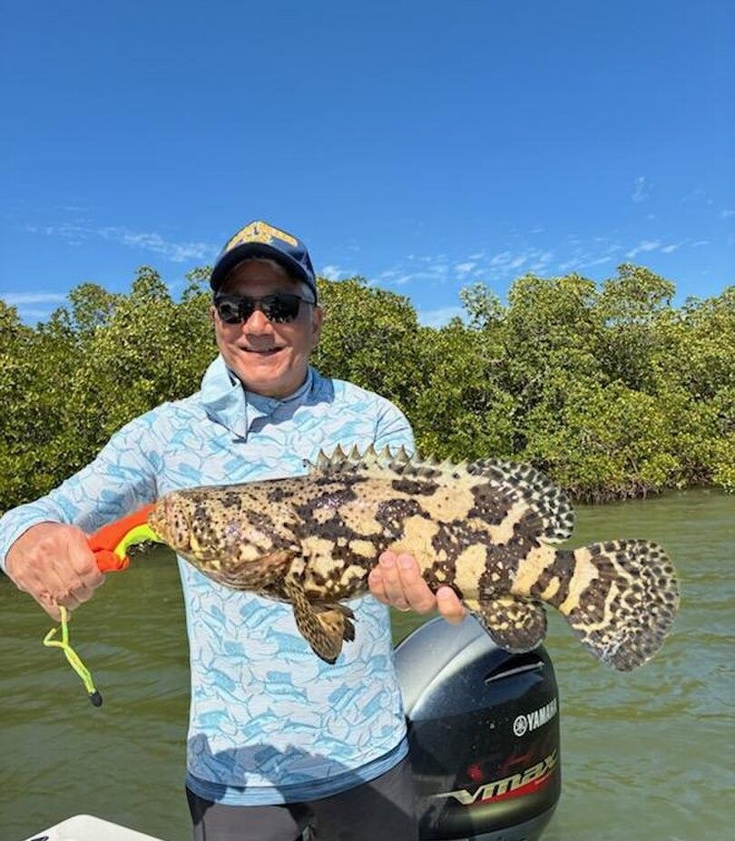 Angler holding caught Goliath Grouper on fishing boat with mangroves in background