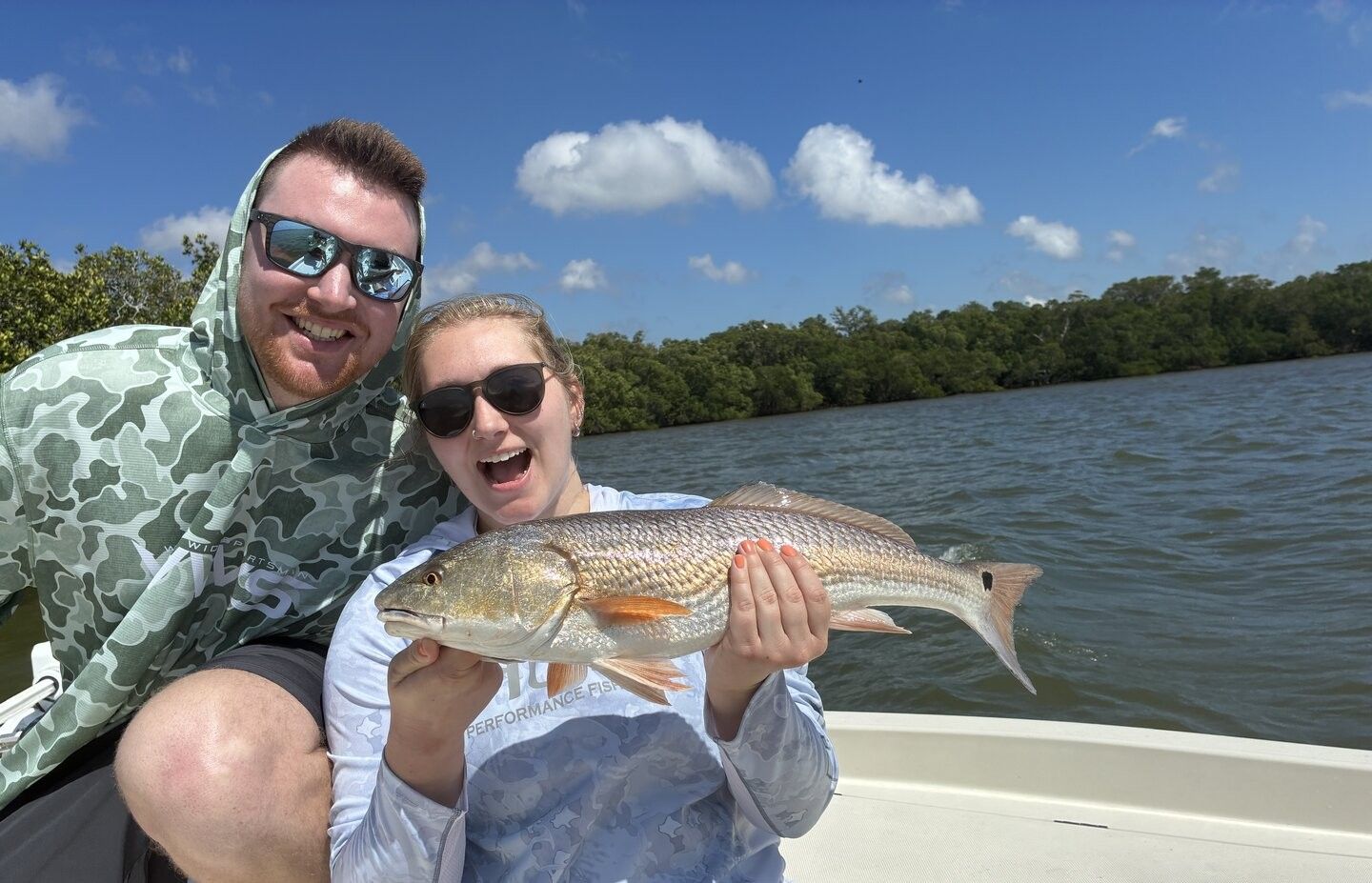Two anglers holding a caught redfish on a boat with water and trees in background