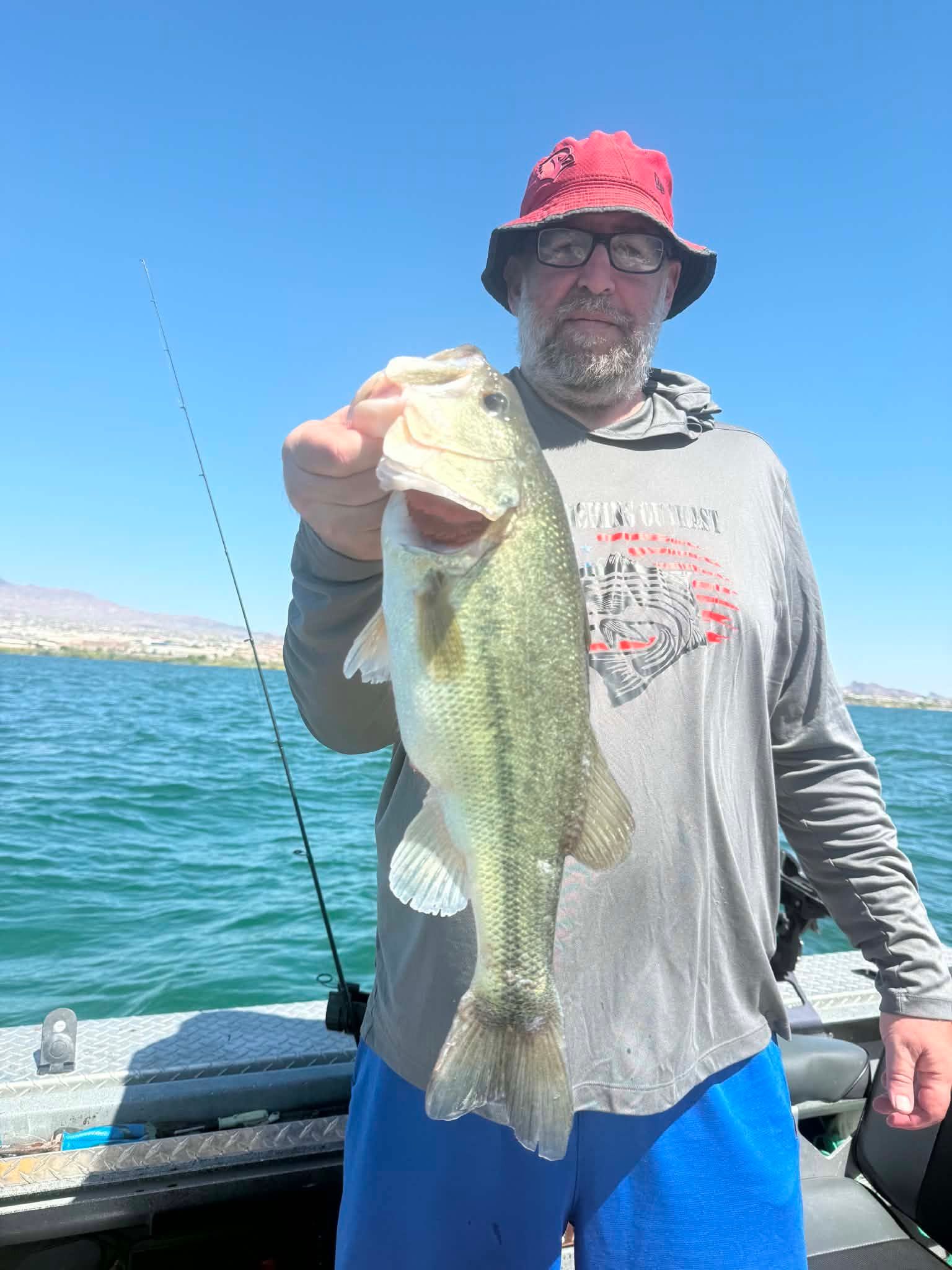 Large bass fish being held up on fishing boat with clear blue water and mountains in background