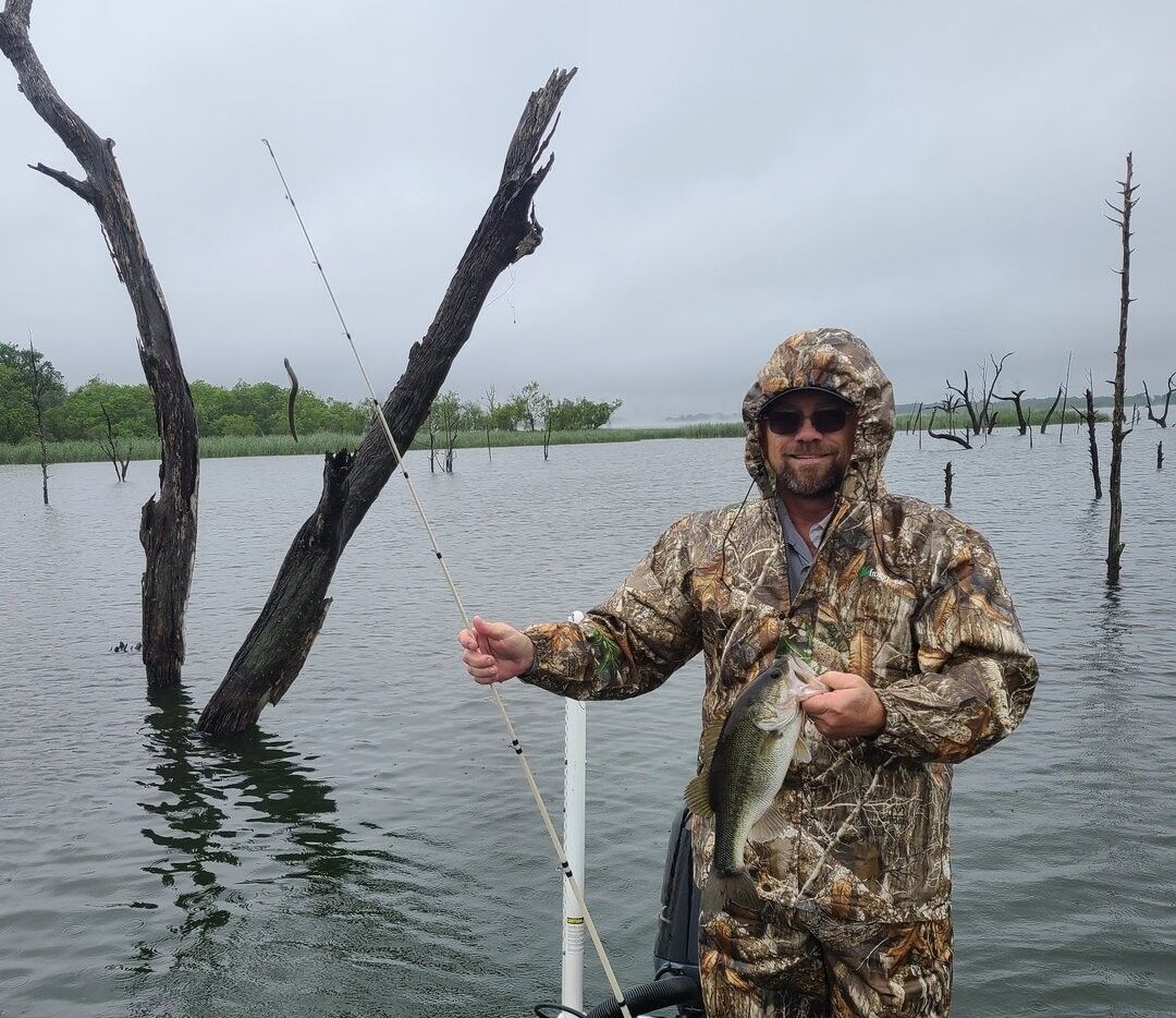 Angler holding caught bass fish on lake with dead trees in background