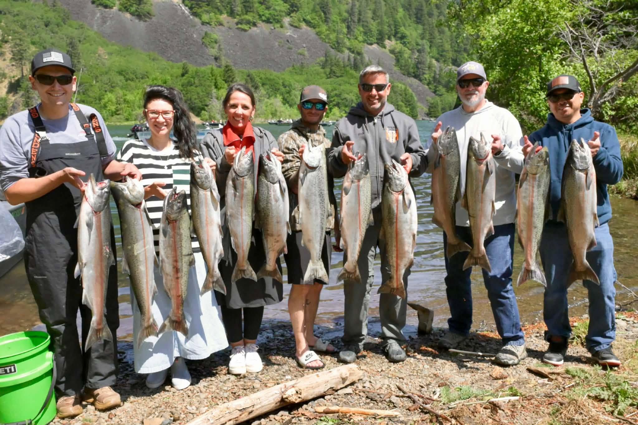 Group of anglers displaying their catch of Chinook and Coho salmon by a river in an outdoor mountain setting