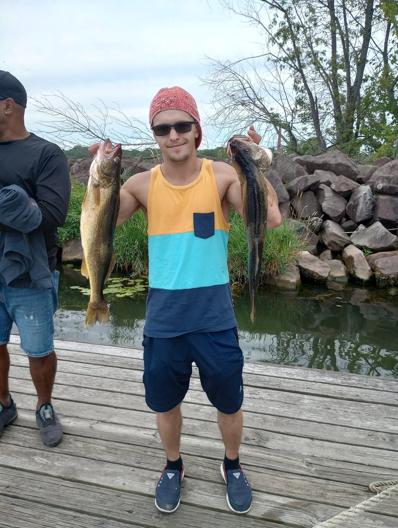 Angler holding two freshly caught sauger fish on wooden dock near water with rocks in background