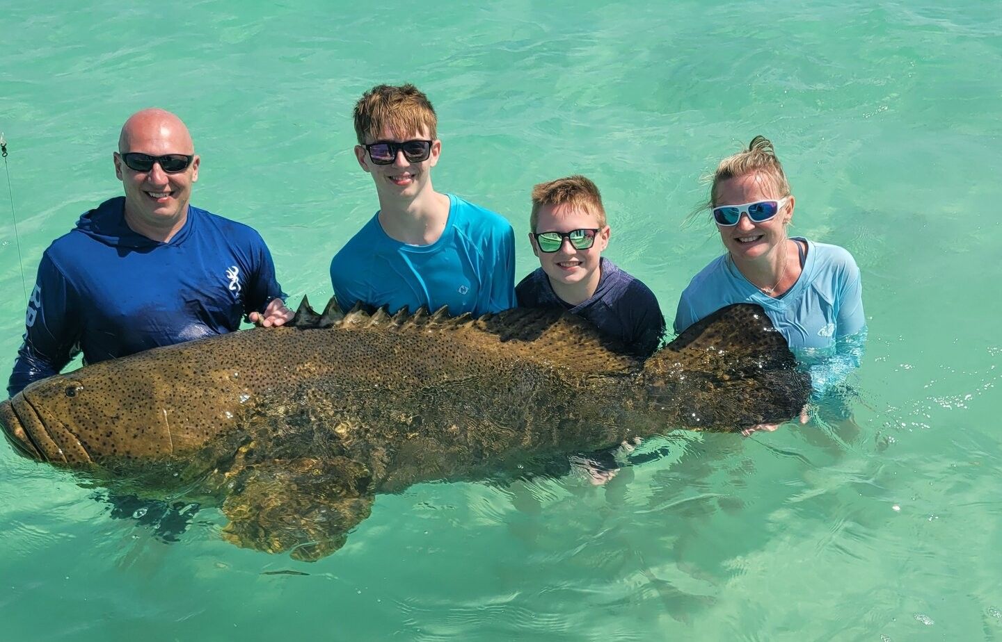 Four people in clear turquoise water holding a massive Goliath Grouper during fishing excursion
