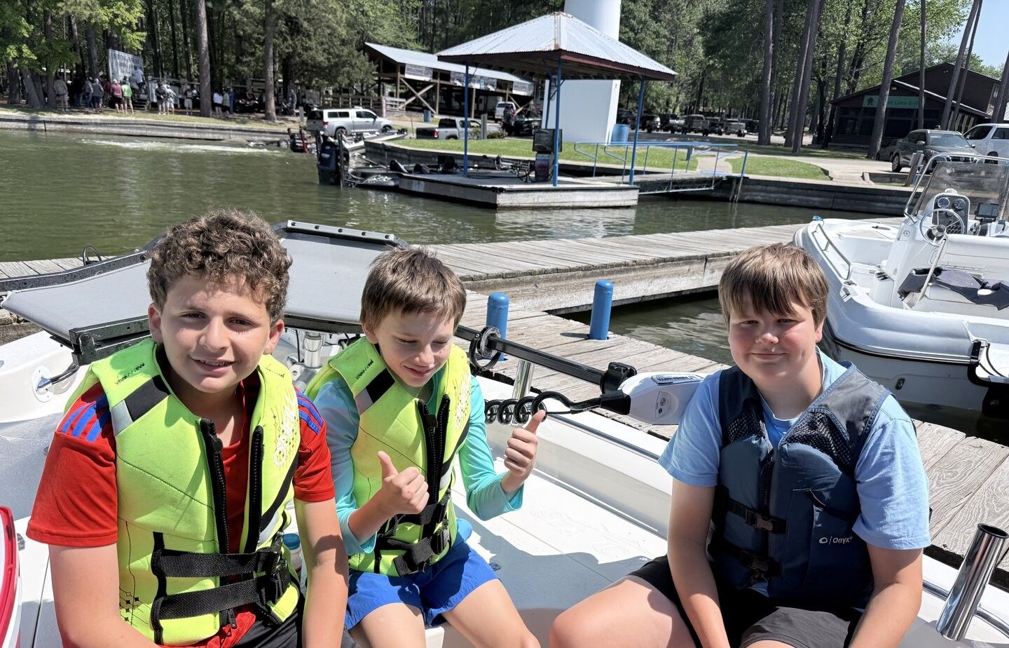 Three young people wearing life jackets sitting on a boat at a marina dock with other boats and trees in the background
