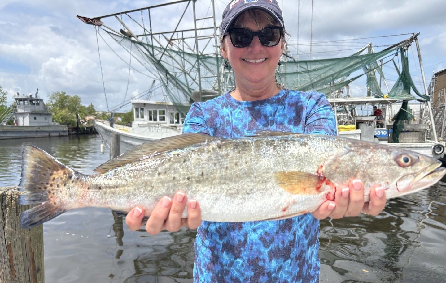 Angler holding freshly caught speckled trout at marina dock with fishing boats in background