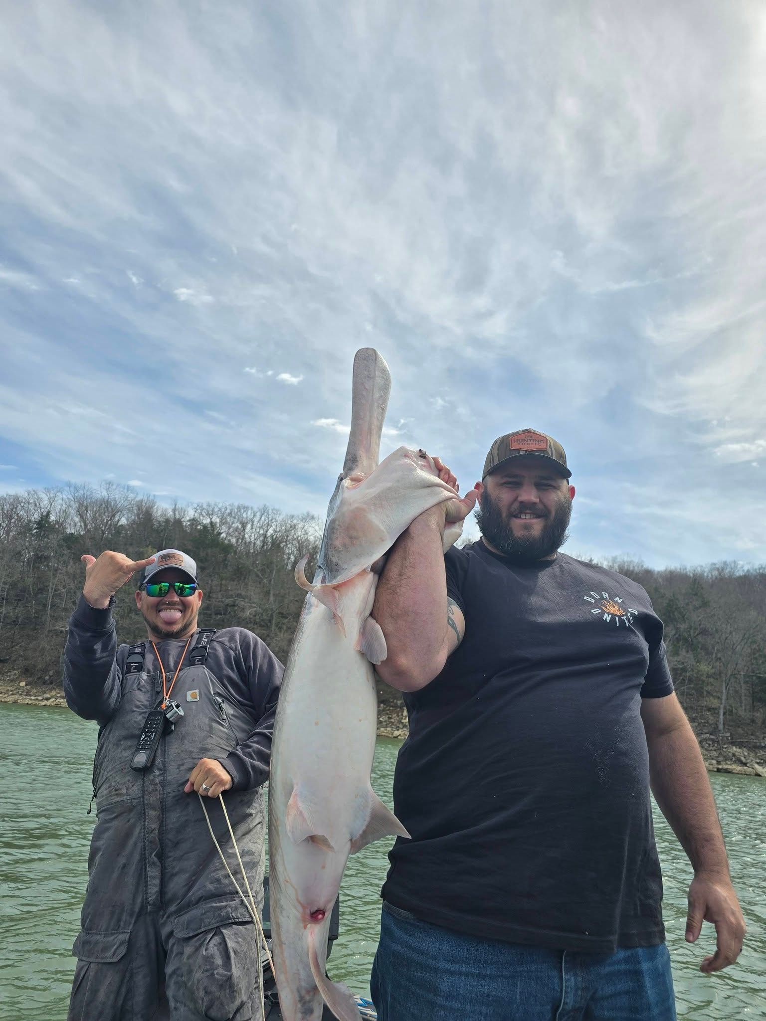 Two anglers displaying a large catfish catch by the water