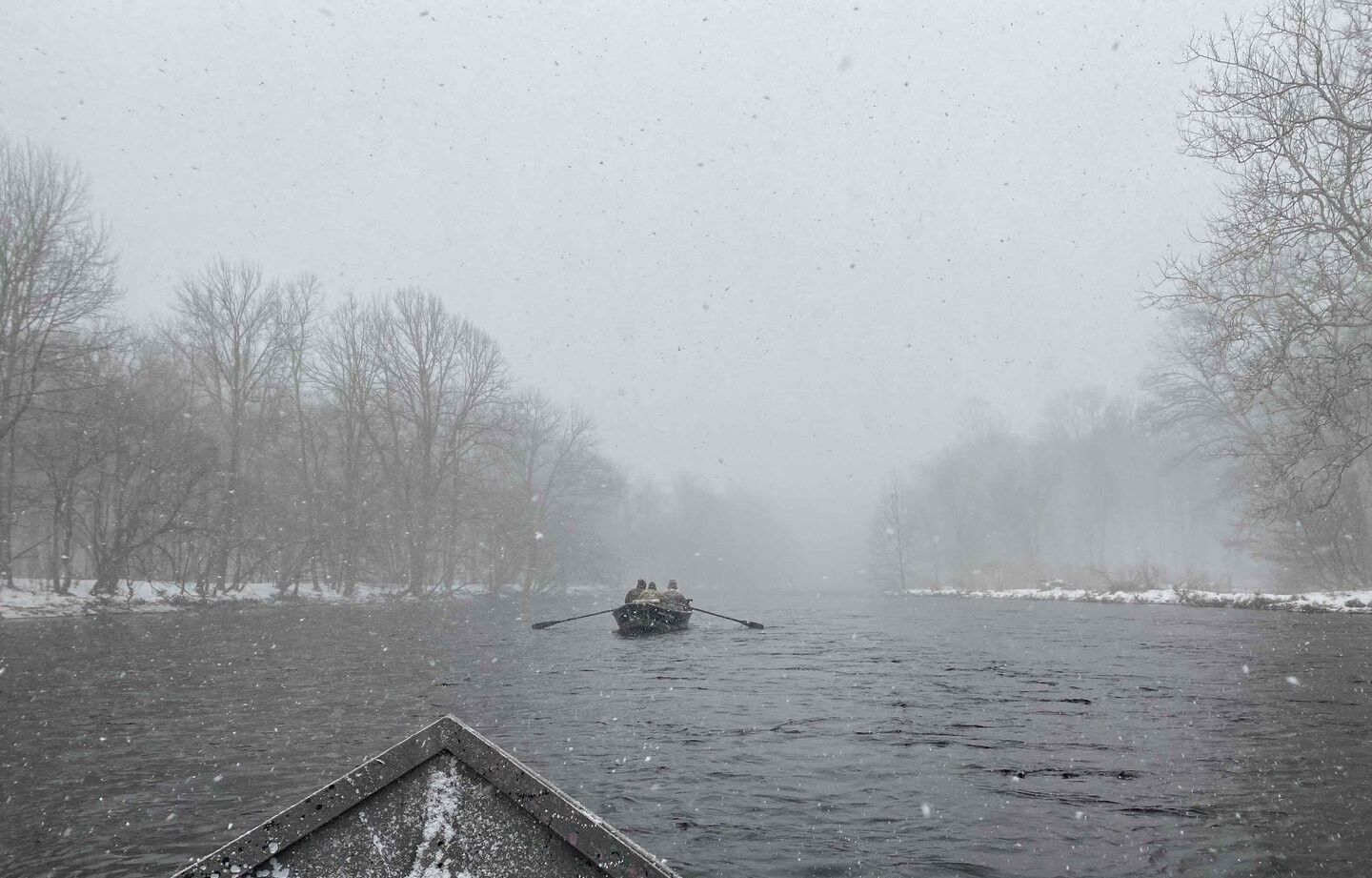 Fishing boat on river during heavy snowfall with bare winter trees along shoreline