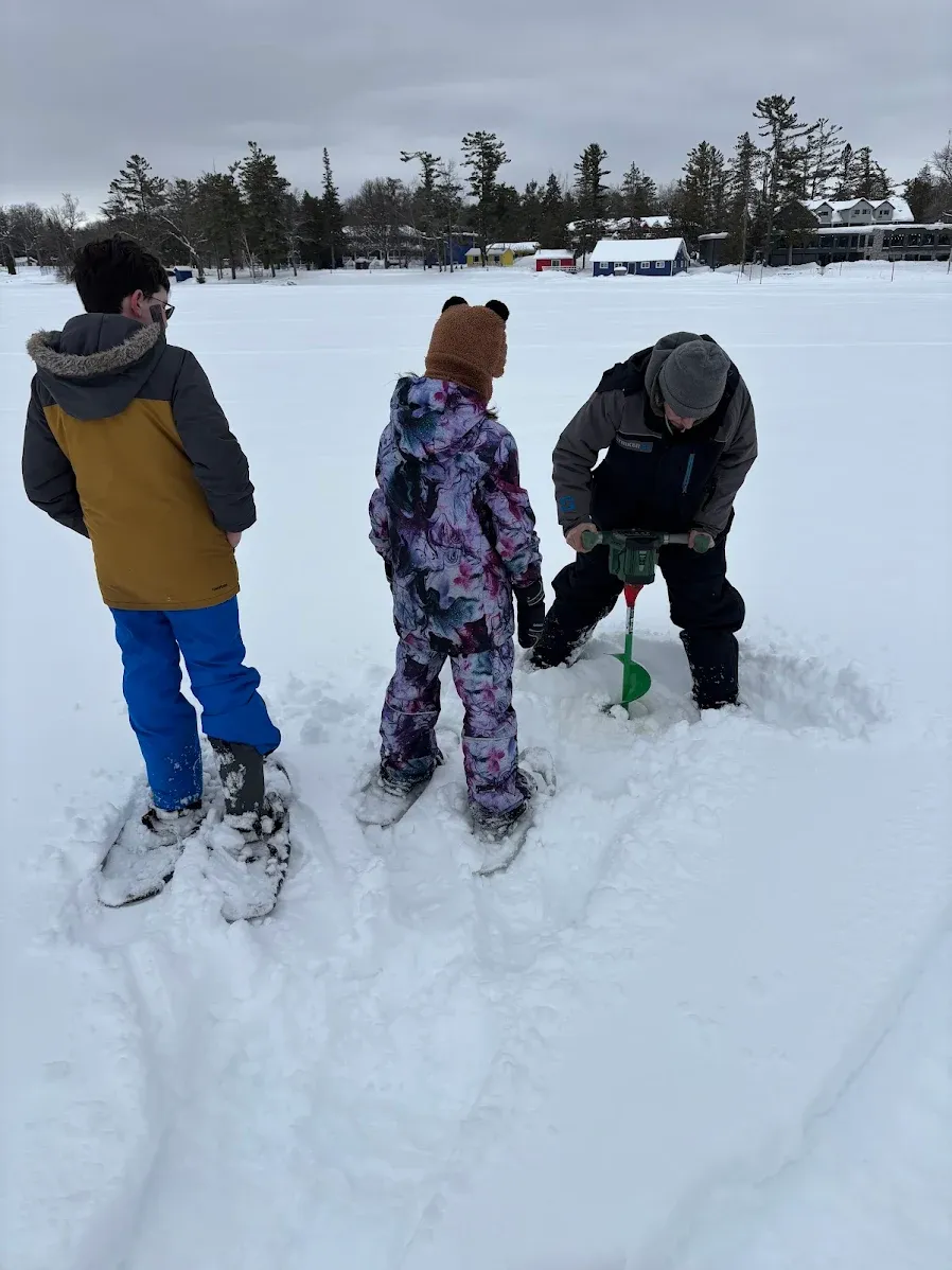 Ice fishing setup with auger drilling hole in frozen lake surrounded by snowy landscape
