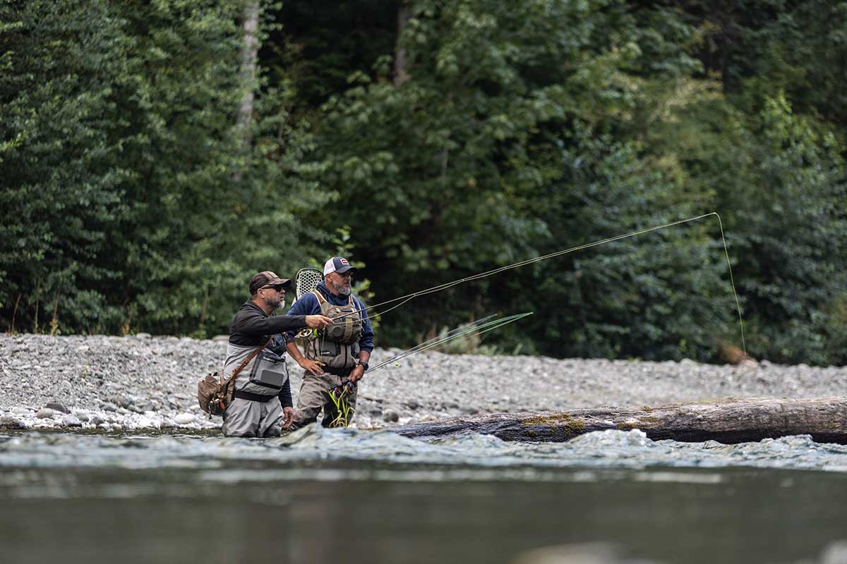 Two anglers fly fishing from rocky shoreline in Gig Harbor Washington with forest backdrop