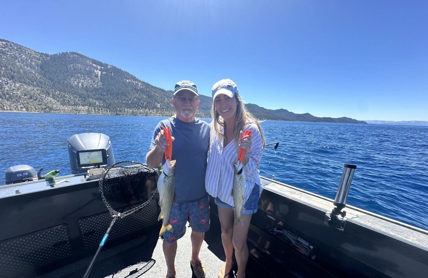 Two anglers displaying caught lake trout on fishing boat at Lake Tahoe Vista CA with mountain backdrop