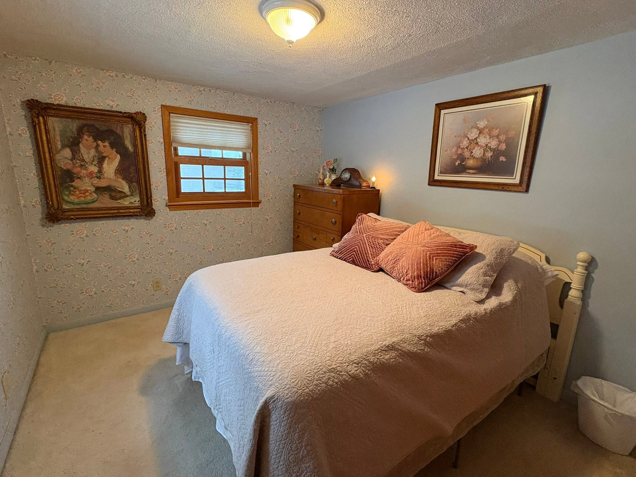 Cozy bedroom with floral wallpaper, wooden dresser, framed artwork, and white bedding in Hale MI