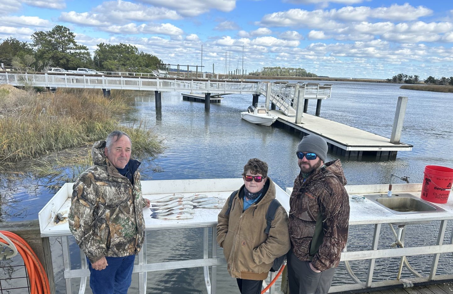 Three anglers displaying their catch of fish on a cleaning table at a Darien GA marina with boats and docks in background