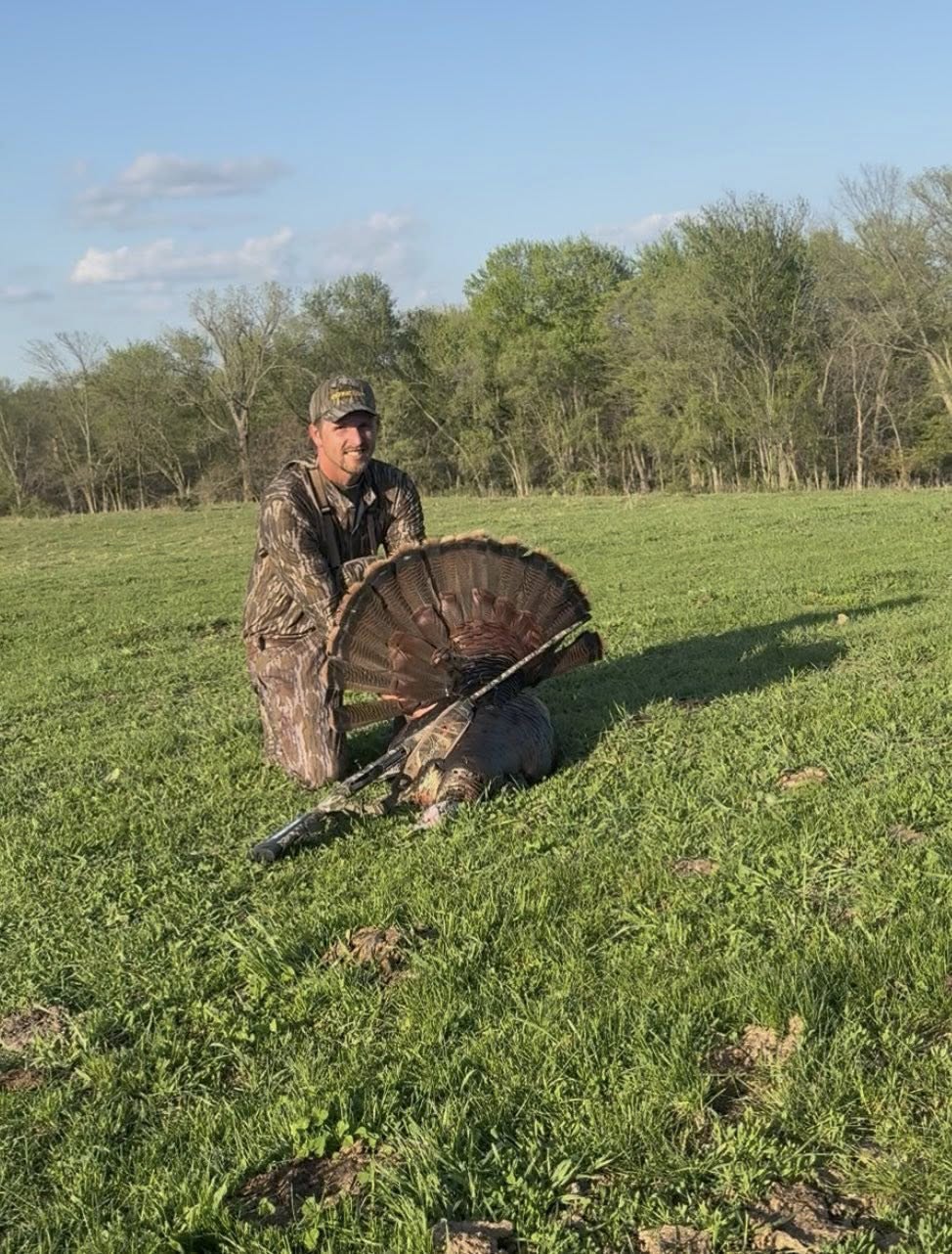 Hunter kneeling in green field displaying harvested wild turkey with fanned tail feathers