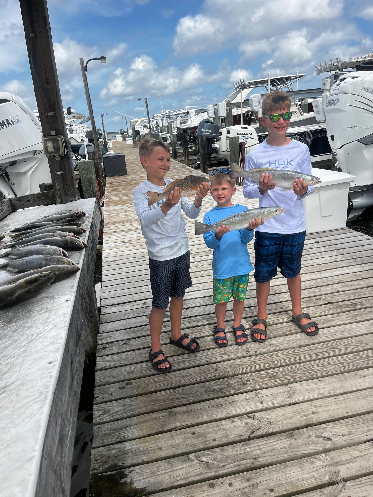 Three young anglers displaying their catch of speckled trout and redfish on a wooden dock at Dauphin Island AL marina