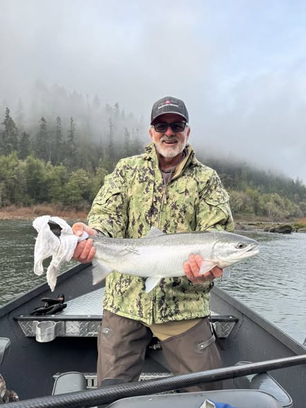 Angler holding large silver fish on boat in Myrtle Creek OR with forested hills in background