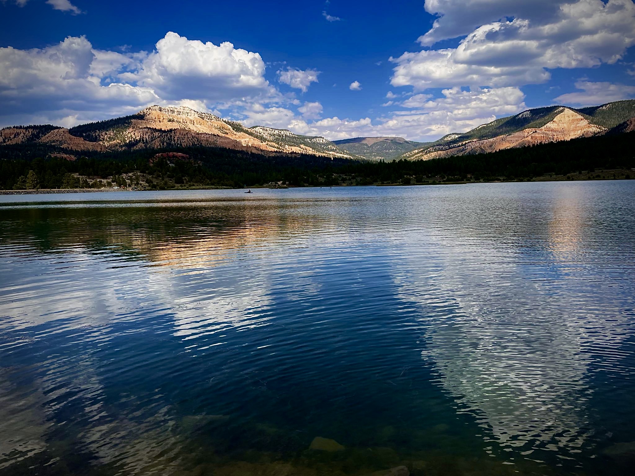 Calm mountain lake surrounded by layered rock formations and forested hills under blue sky with white clouds, perfect for off-road adventure access
