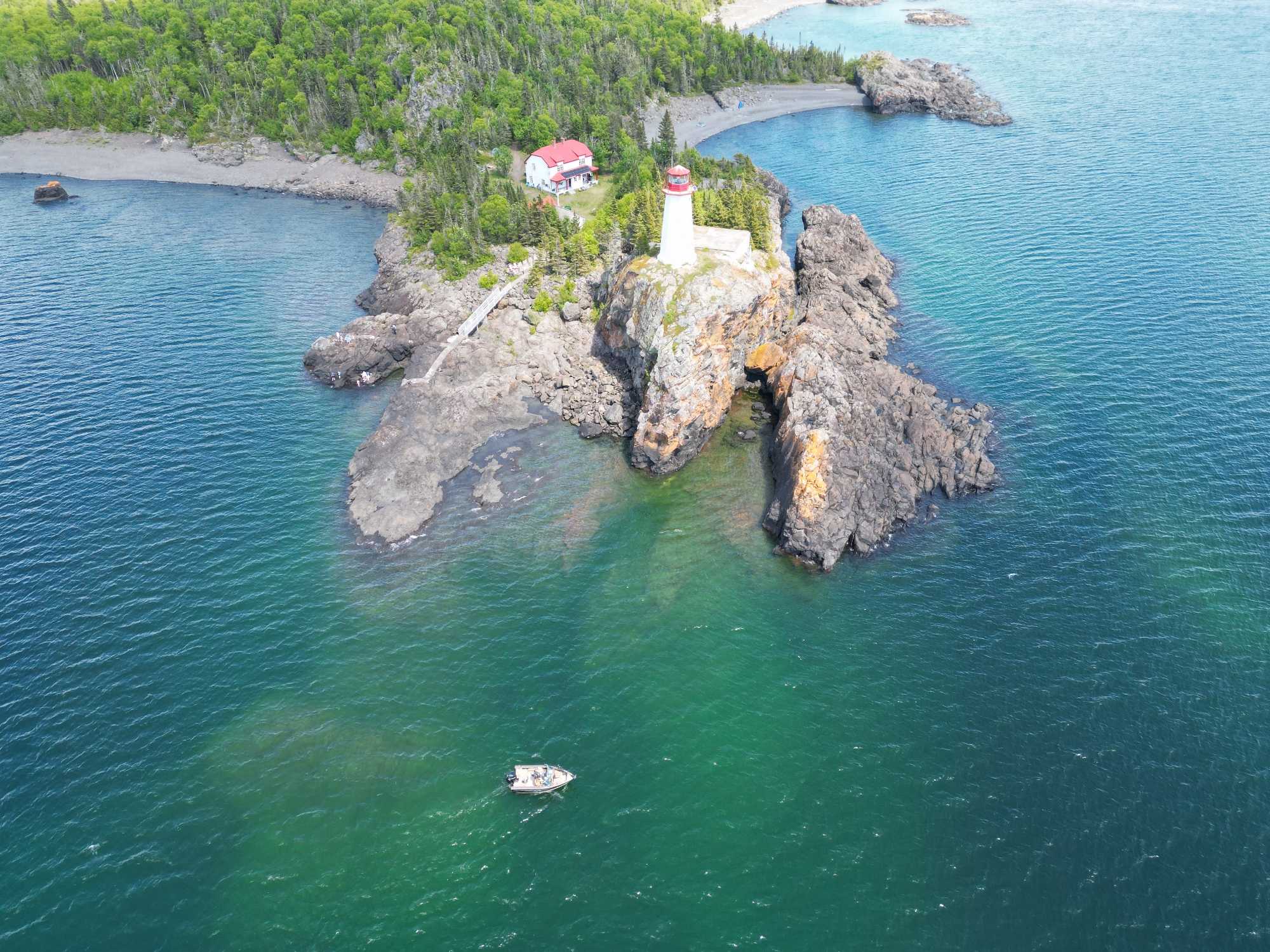 Aerial view of white lighthouse on rocky outcrop surrounded by clear blue-green water near Nipigon ON with small boat nearby