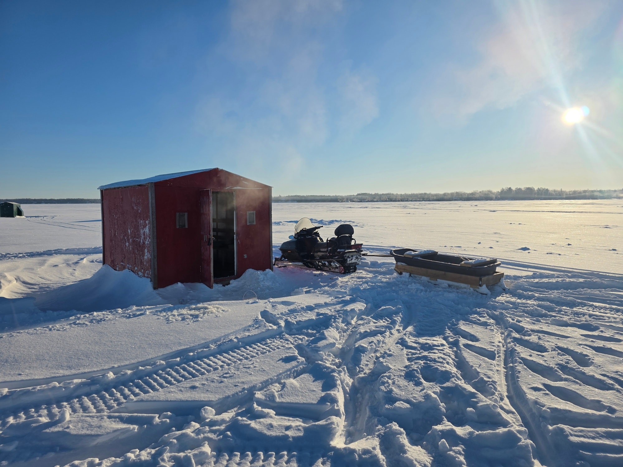 Red ice fishing hut with snowmobile and sled on frozen lake in Gravenhurst ON under sunny winter sky