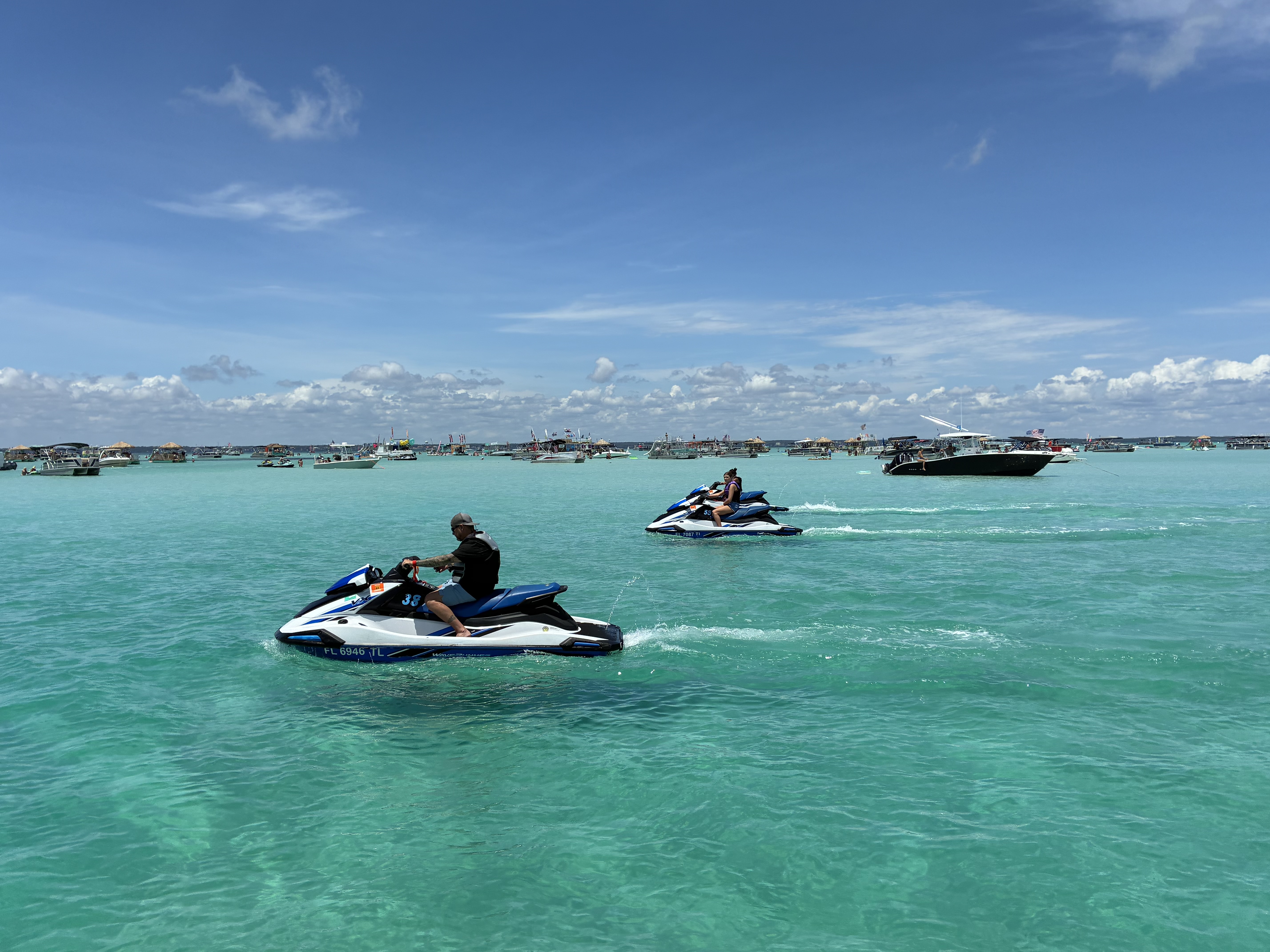 Two jet skiers riding through crystal clear turquoise waters with boats anchored in the background