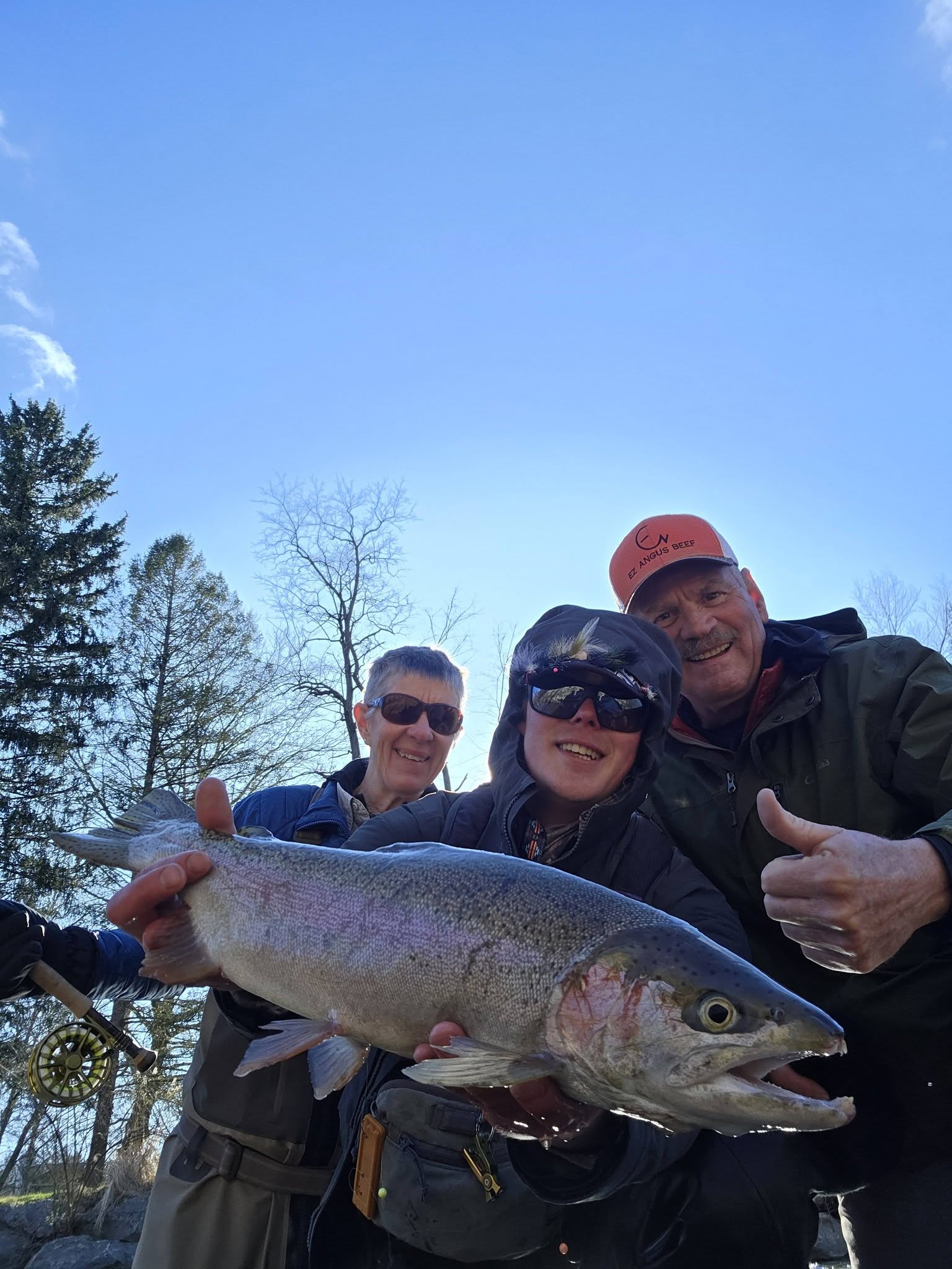 Three anglers holding large rainbow trout catch in Angola NY with trees and clear sky in background